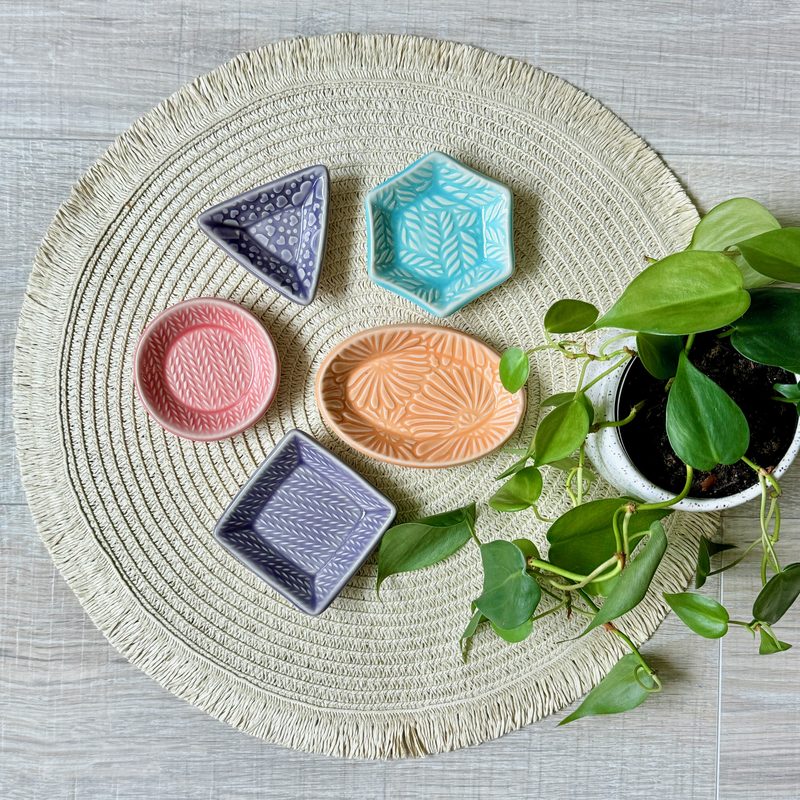 Colorful geometric trinket dishes on woven placemat beside plant.