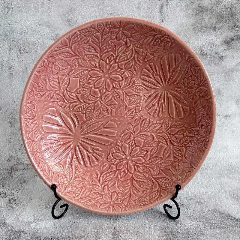 Large round pink ceramic plate with carved flowers and butterflies displayed upright on a stand