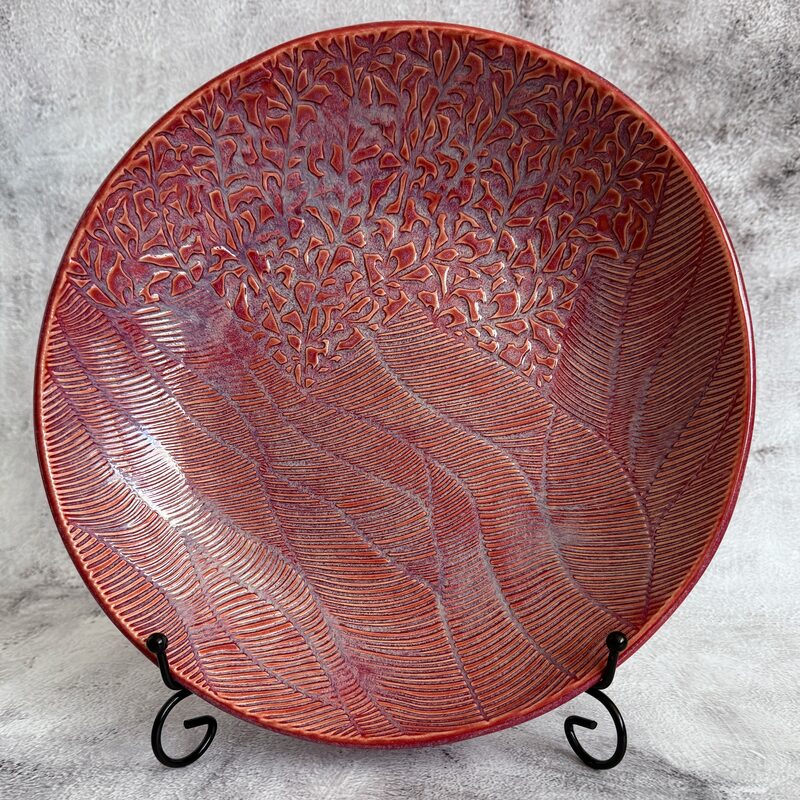 Large round red ceramic platter with carved wave and leaf pattern presented on a display stand