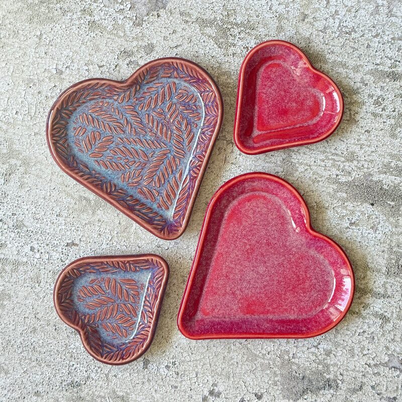 Group of five heart-shaped ceramic trays in red and blue leaf and solid glazes styled on a concrete surface