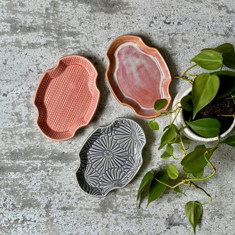 Three ornate-edge ceramic trays in soft pink, peach and gray floral patterns displayed beside a potted plant