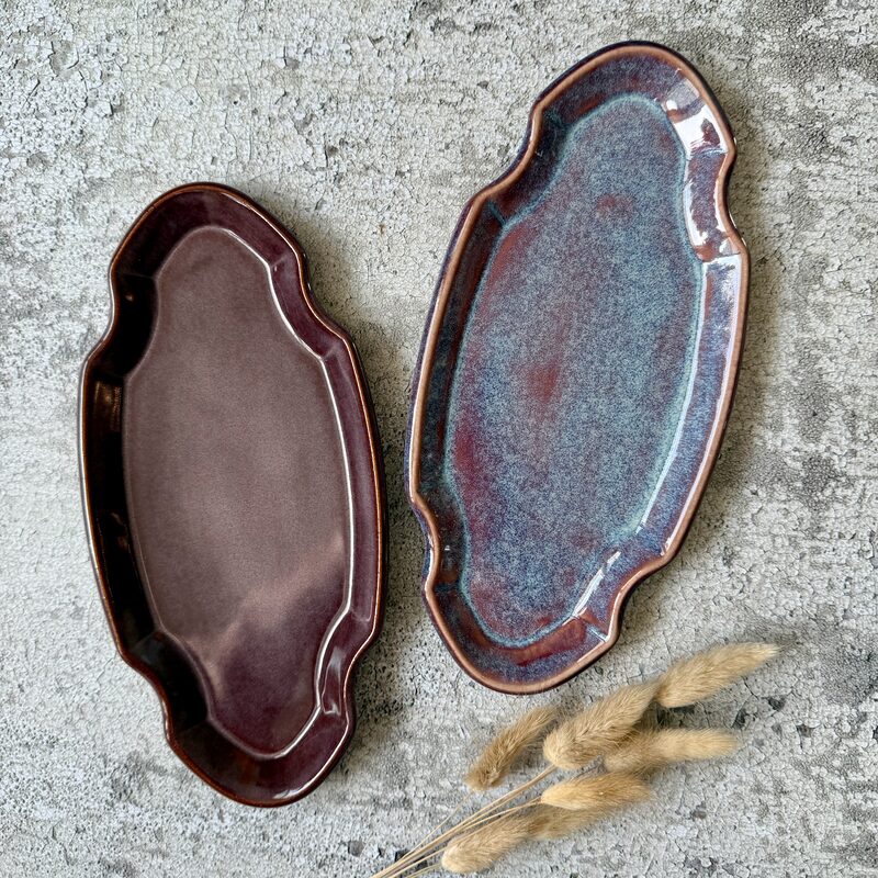 Pair of long oval ceramic serving platters in plum and blue-red glazes surrounded by dried grasses